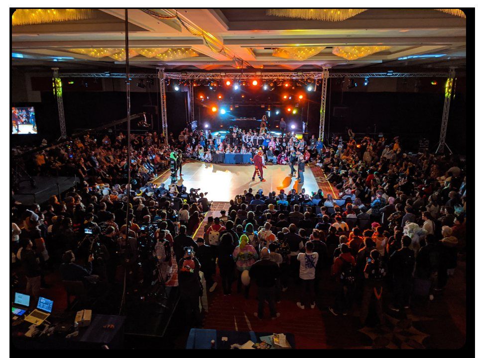 A packed convention hall seen from above, with the audience gathered around a brightly lit stage.
